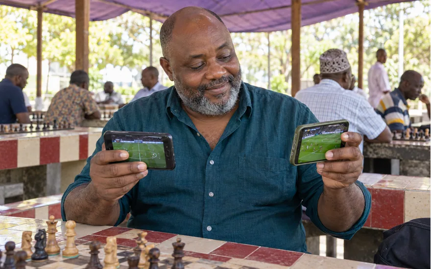Adult Tanzania reader comparing two sports phones at a shaded outdoor game shelter