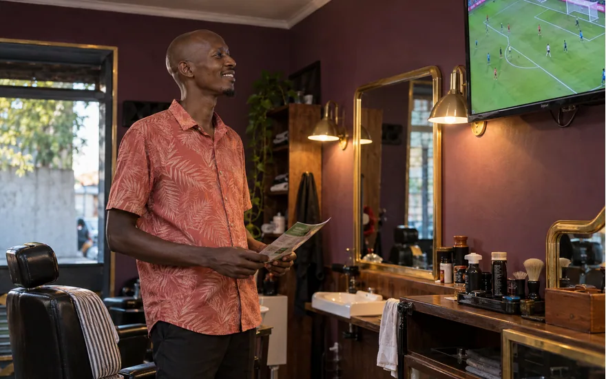 Adult Tanzania reader holding a promotion sheet beside a football screen in an after-hours barber shop